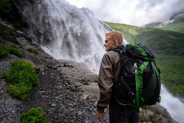 Mann wandert mit Rucksack und blickt auf Wasserfall. Travel Lifestyle Adventure Konzept, Aktivurlaub in wilder Harmonie mit der Natur. Nordkaukasus, Dombai, Russland. - Foto, Bild