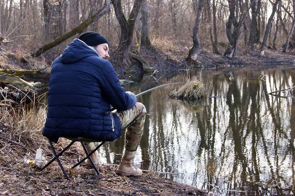 Rybářská prut v rukou rybáře na jezeře. Mladý muž sedí na židli s otočnou tyčí na pozadí vody. - Fotografie, Obrázek