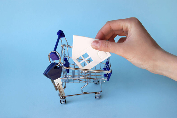 A hand puts a paper white house in a mini shopping trolley on a blue background. The concept of buying or renting a house, apartment, cottage and much more. - Photo, Image
