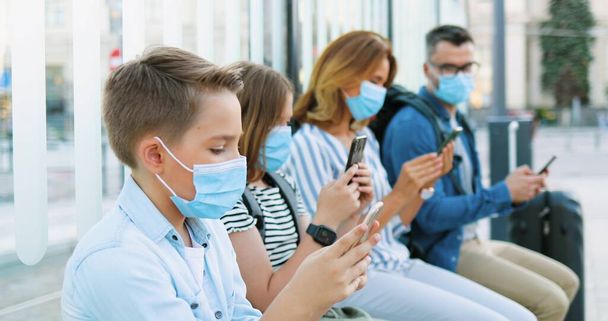 Caucasian family with children in medical masks sitting at bus stop, waiting for transport. Boy and girl with mother and father using phones. Kids and adults tapping and scrolling on smartphones. - Фото, изображение