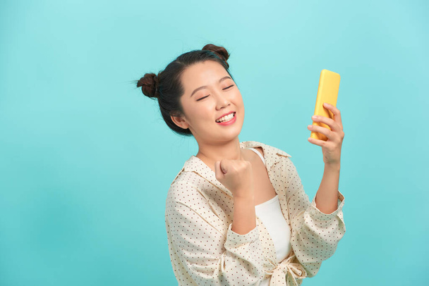 Fröhliche Asiatin steht isoliert vor blauem Hintergrund, hält Handy in der Hand, feiert - Foto, Bild