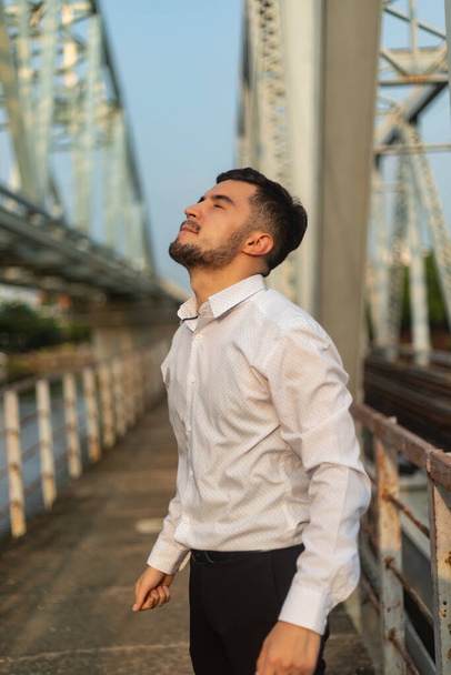 A vertical shot of a young Caucasian man with a white shirt standing on a bridge - Fotoğraf, Görsel