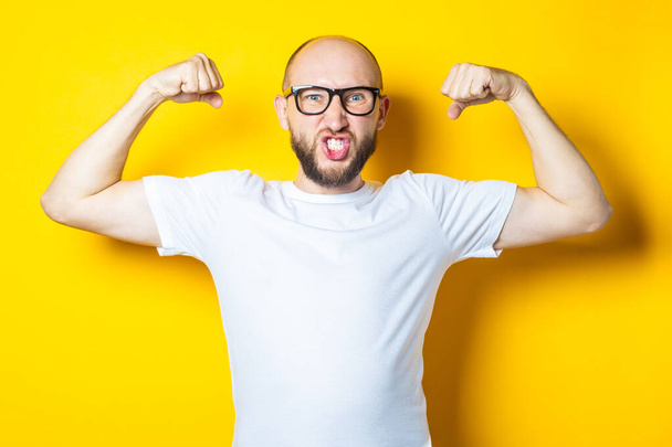 Kale jongeman met een baard toont spieren, kracht op een gele achtergrond - Foto, afbeelding