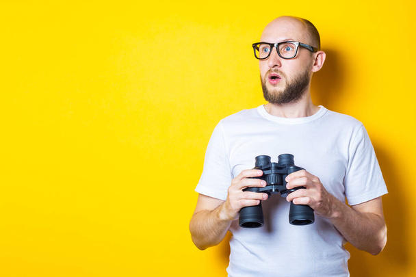 Surprised shocked bearded young man looking to the side while holding binoculars on yellow background. - Foto, immagini