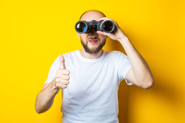 Smiling bald young man with a beard looks through binoculars and shows a thumb up on a yellow background. - Fotó, kép