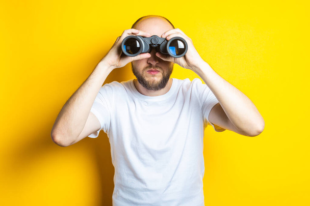Young man with a beard looking through binoculars on a yellow background - Foto, Imagen