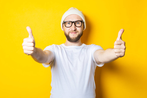 Smiling affable young guy with a beard in a cap and glasses shows a gesture with his hands, thumbs up, class on a yellow background - Photo, Image