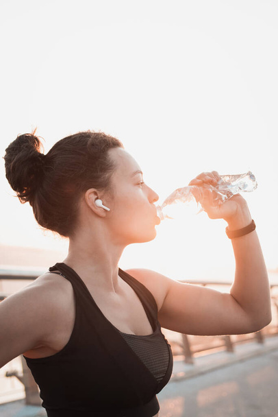 Athletic woman runner in modern headphones drinking water and taking rest break after working out,exercising, standing outdoors on city bridge in the morning sunrise background.Vertical.Water balance. - Фото, изображение