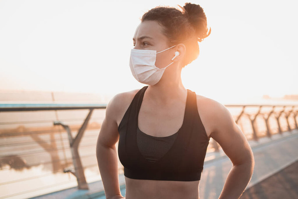 Sport during quarantine,coronavirus,covid-19 concept.Young athletic woman runner wearing medical protective mask taking a rest after training jogging exercise outdoor on city bridge in the morning. - Photo, Image