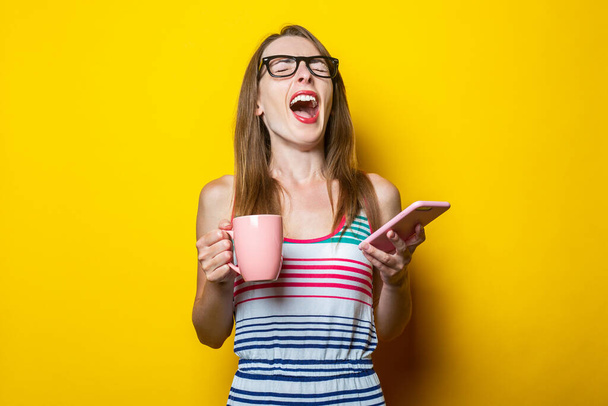 Молода дівчина кричить, тримаючи телефон і чашку з кавою на жовтому фоні
 - Фото, зображення