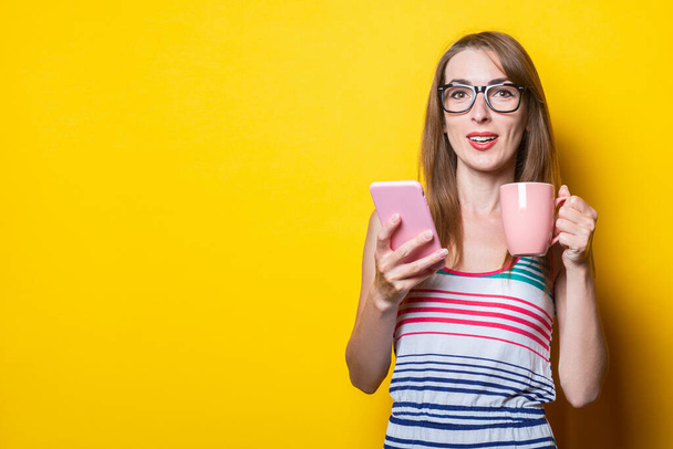 Young woman in striped dress drinks coffee with phone on yellow background - Photo, Image