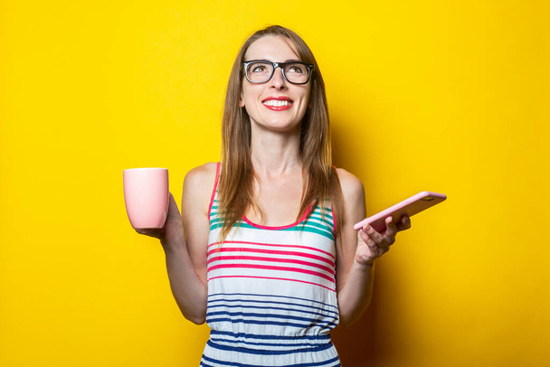 Beautiful young smiling woman looks up holding a phone and a cup with coffee on a yellow background. - Photo, Image