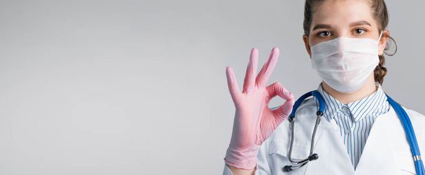 Beyaz önlüklü, tıbbi maskeli ve pembe steril eldivenli güzel bir kadın doktor gri arka planda sorun olmadığını gösteriyor. Sağlıklı insan kavramı mı yoksa bir doktora güvenmek mi? - Fotoğraf, Görsel