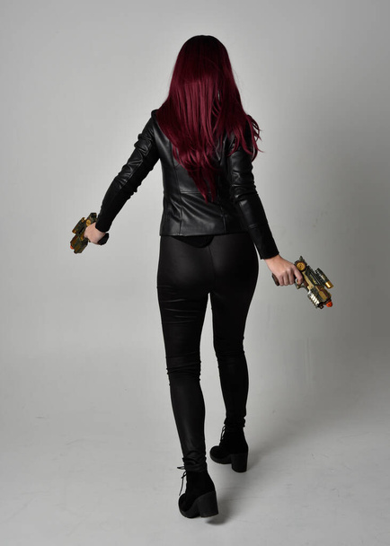 Full length portrait of  girl with red hair wearing black leather jacket, pants and boots. Standing pose holding a gun with back to the camera, isolated against a grey studio background. - Photo, Image
