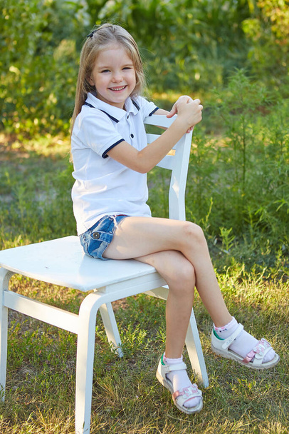 Little charming girl sitting on a white chair in the summer garden - Zdjęcie, obraz