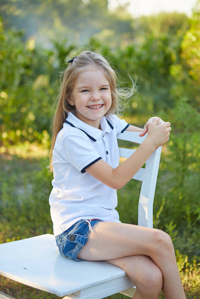Little charming girl sitting on a white chair in the summer garden - Foto, immagini