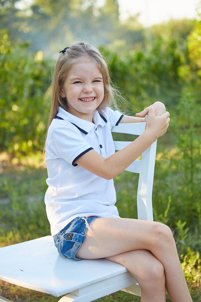 Little charming girl sitting on a white chair in the summer garden - Photo, Image