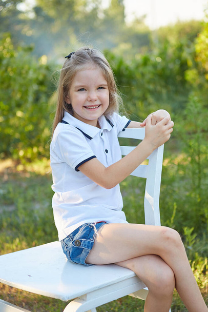 Little charming girl sitting on a white chair in the summer garden - Fotoğraf, Görsel
