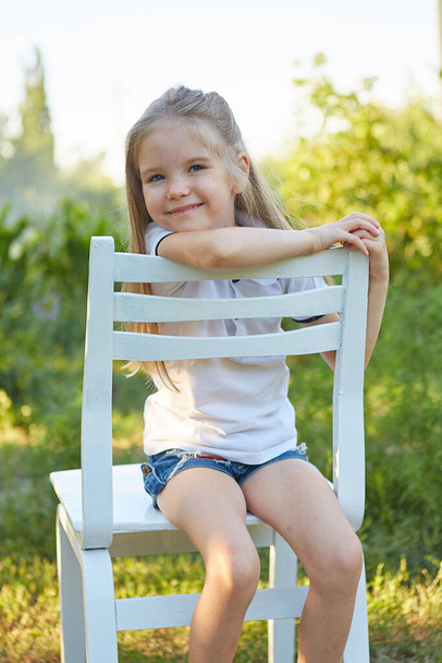 Little charming girl sitting on a white chair in the summer garden - Photo, image