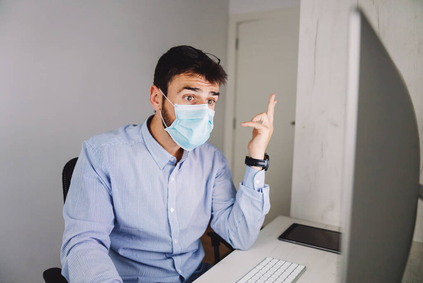 Νεαρός αφοσιωμένος επιχειρηματίας με μάσκα προσώπου να κάθεται στο γραφείο του και να χρησιμοποιεί υπολογιστή για δουλειά κατά τη διάρκεια της επιδημίας του ιού Κορόνα. - Φωτογραφία, εικόνα