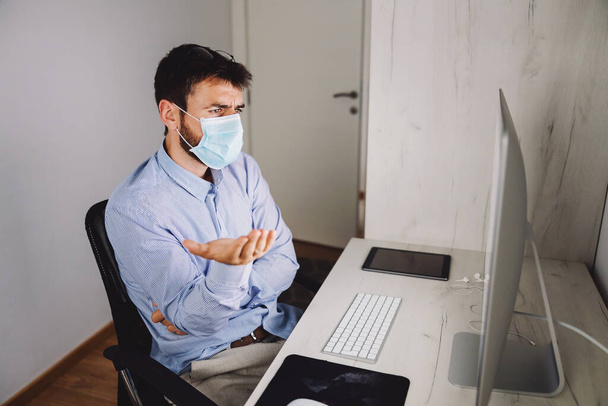 Молодой бизнесмен в маске для лица смотрит на монитор компьютера и ищет решение, сидя в своем офисе во время вспышки коронного вируса. - Фото, изображение