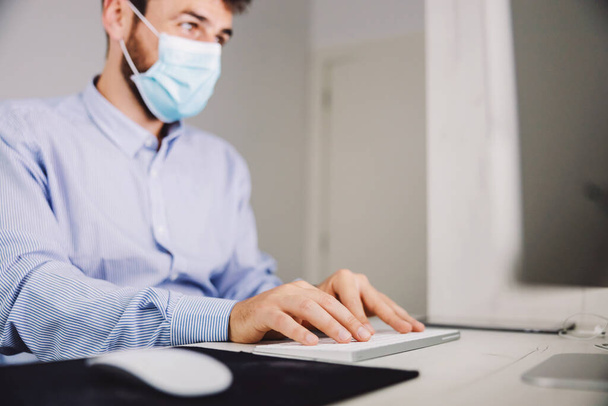 Closeup of young hardworking businessman with face mask on sitting in his office and typing report during covid 19 outbreak. - Fotografie, Obrázek
