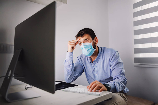 Młody atrakcyjny pracownik nerwowy z maską na twarzy siedzi w swoim biurze i czyta ważne e-maile od klienta podczas wybuchu choroby. - Zdjęcie, obraz