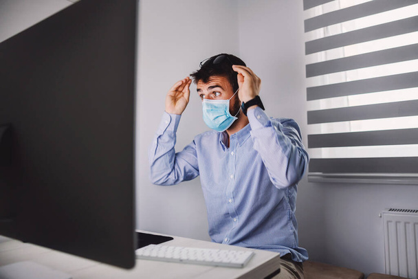 Młody, oddany biznesmen z maską na twarzy, siedzący w biurze podczas epidemii wirusa korony i patrzący na komputer. - Zdjęcie, obraz