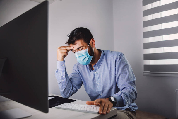 Przystojny pracownik siedzi w biurze z maską na twarzy i trzyma głowę podczas epidemii wirusa korony. - Zdjęcie, obraz