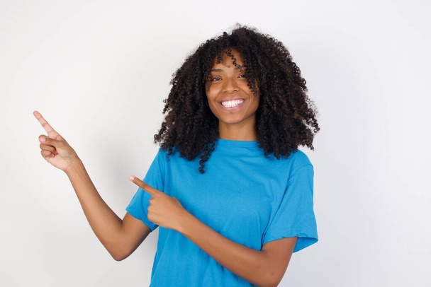 bonita jovem mulher africana com cabelo encaracolado vestindo camisa azul casual em pé contra fundo branco indicando com indicador espaço vazio mostrando os melhores preços baixos
  - Foto, Imagem