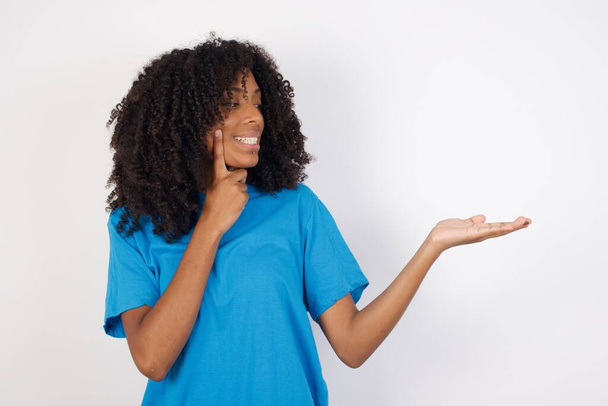 Mujer africana joven divertida con el pelo rizado con camisa azul casual de pie sobre fondo blanco con el pelo corto sosteniendo palma abierta nuevo producto. Quiero comprarlo.!  - Foto, imagen