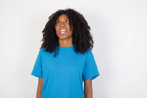 Kıvırcık saçlı, günlük mavi gömlekli, beyaz arka planda duran, yüzünü ekşiten ve dilini gösteren Afrikalı genç bir kadın. Komik ve çılgın olmak - Fotoğraf, Görsel