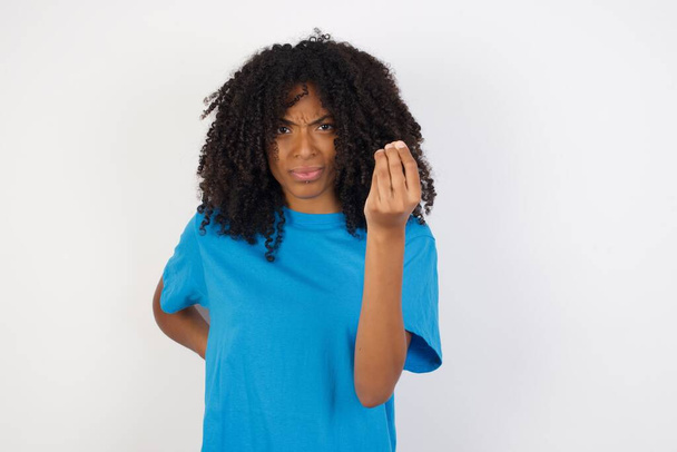 Молодая африканская женщина с вьющимися волосами в повседневной синей рубашке стоит на белом фоне сердито жестикулируя типичным итальянским жестом с рукой, глядя в камеру - Фото, изображение