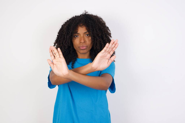 Mujer africana joven con el pelo rizado con camisa azul casual de pie sobre fondo blanco tiene expresión de rechazo cruzando brazos y palmas haciendo signo negativo, cara enojada. - Foto, imagen