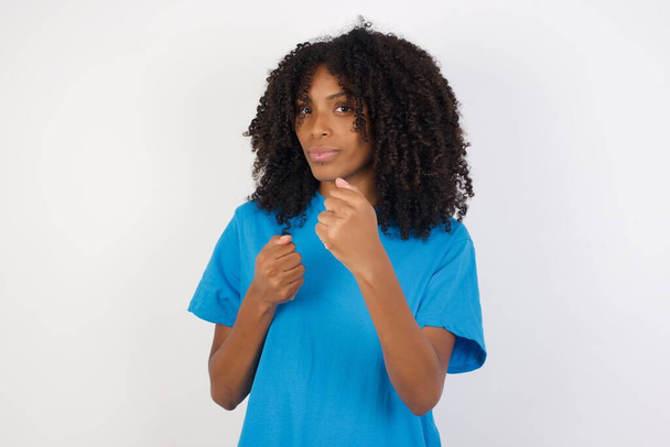 Молода африканська жінка з кучерявим волоссям в синій сорочці, що стоїть на білому тлі Готовий боротися з жестом захисту кулака, розлючене і засмучене обличчя, боїться проблем
. - Фото, зображення