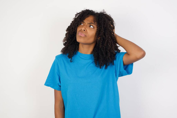 Junge Afrikanerin mit lockigem Haar, lässig blauem Hemd vor weißem Hintergrund, verwirrt und wundert sich über Fragen. Unsicher im Zweifel, mit der Hand auf dem Kopf denkend. Umfassendes Konzept. - Foto, Bild