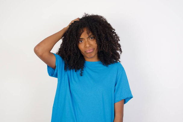 Молодая африканская женщина с вьющимися волосами, в повседневной синей рубашке, стоящей на белом фоне, смущается и задается вопросом. Держась за голову, неуверенная в себе. Комплексная концепция. - Фото, изображение