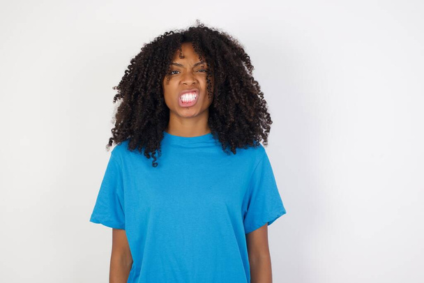 Портрет сердитой молодой африканской женщины с вьющимися волосами, носящей случайную голубую рубашку, стоящую на белом фоне, держит зубы сжатыми, хмурится лицом в неудовлетворенности, раздражается из-за большого количества обязанностей. Разъяренная девушка - Фото, изображение
