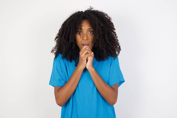 Mujer africana joven con el pelo rizado con camisa azul casual de pie sobre fondo blanco rezando por la suerte tiene las manos cruzadas cerca de la cara, asombrado y la boca abierta mirando hacia el frente. - Foto, Imagen