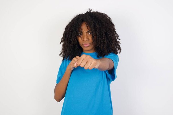 Retrato de mulher africana jovem forte e determinada com cabelo encaracolado vestindo camisa azul casual em pé contra fundo branco perfurando o ar com punho e olhando com confiança para a câmera, luta masculina, espírito de luta
.  - Foto, Imagem