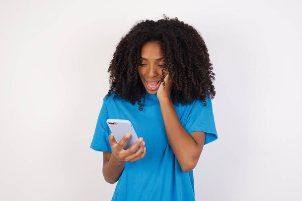 Close-up portrait of Young african woman with curly hair wearing casual blue shirt standing against white background holding in hands cell reading sms using new app 5g  - Foto, Imagem