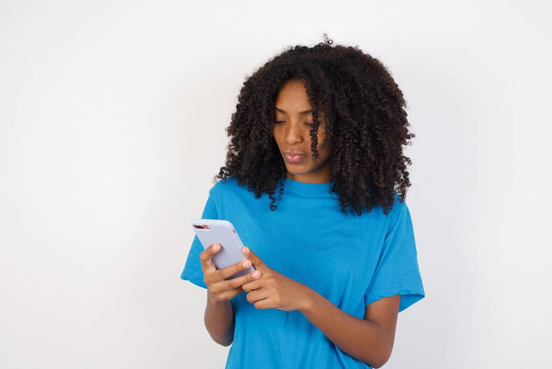 Portrait de fou impressionné Jeune femme africaine aux cheveux bouclés portant une chemise bleue décontractée debout sur fond blanc utiliser téléphone portable lit des nouvelles incroyables sur les médias sociaux information regard, cris wow usure omg  - Photo, image
