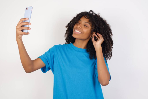 Foto der charmanten jungen Afrikanerin mit lockigem Haar, die lässig blaues Hemd vor weißem Hintergrund trägt, lächelt und ein Selfie macht, um es in ihren sozialen Medien zu posten. - Foto, Bild
