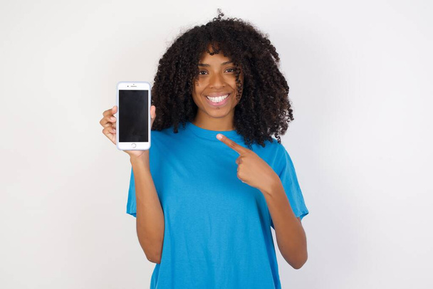 vrolijk aantrekkelijk Jonge Afrikaanse vrouw met krullend haar dragen casual blauw shirt staan tegen een witte achtergrond met telefoon in de hand nieuw model van appel telefoon gewoon kijken naar de prijs - Foto, afbeelding