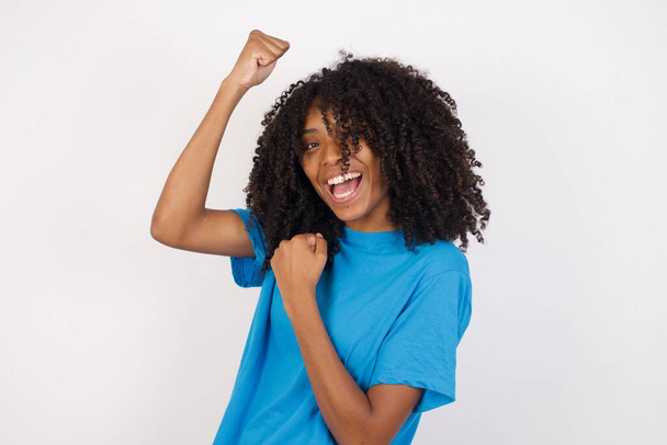 Profiel foto van opgewonden Jonge Afrikaanse vrouw met krullend haar dragen casual blauw shirt staan tegen witte achtergrond verhogen vuisten vieren zwarte vrijdag winkelen  - Foto, afbeelding