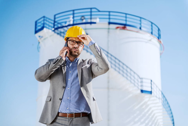 Serious Caucasian handsome businessman in suit and with helmet on head talking on smart phone while standing outdoors at refinery. In background is tank with oil. - Photo, Image