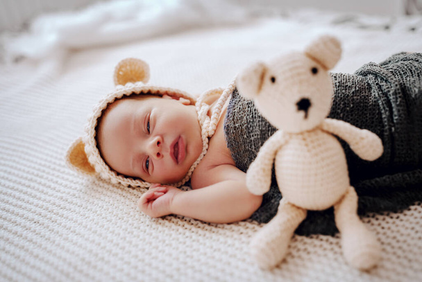 Vista lateral de un hermoso bebé recién nacido envuelto en una bufanda de lana y con gorra en la cabeza durmiendo al lado de un juguete. - Foto, Imagen