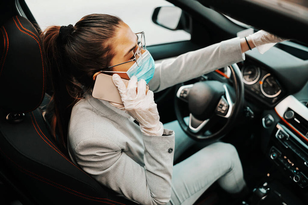 Młoda atrakcyjna brunetka z maską na twarzy z gumowymi rękawiczkami siedząca w samochodzie i dzwoniąca podczas wybuchu epidemii. - Zdjęcie, obraz