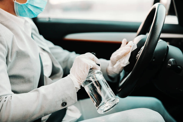 lähikuva naisesta, jolla on kasvonaamio ja kumihanskat ruiskuttamalla ohjauspyörää alkoholilla ja desinfioimalla autonsa koronaviruksen puhjetessa. - Valokuva, kuva