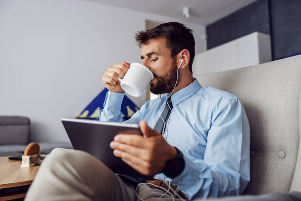 Knappe bebaarde zakenman die koffie drinkt en tablet gebruikt terwijl hij thuis in de stoel zit. - Foto, afbeelding